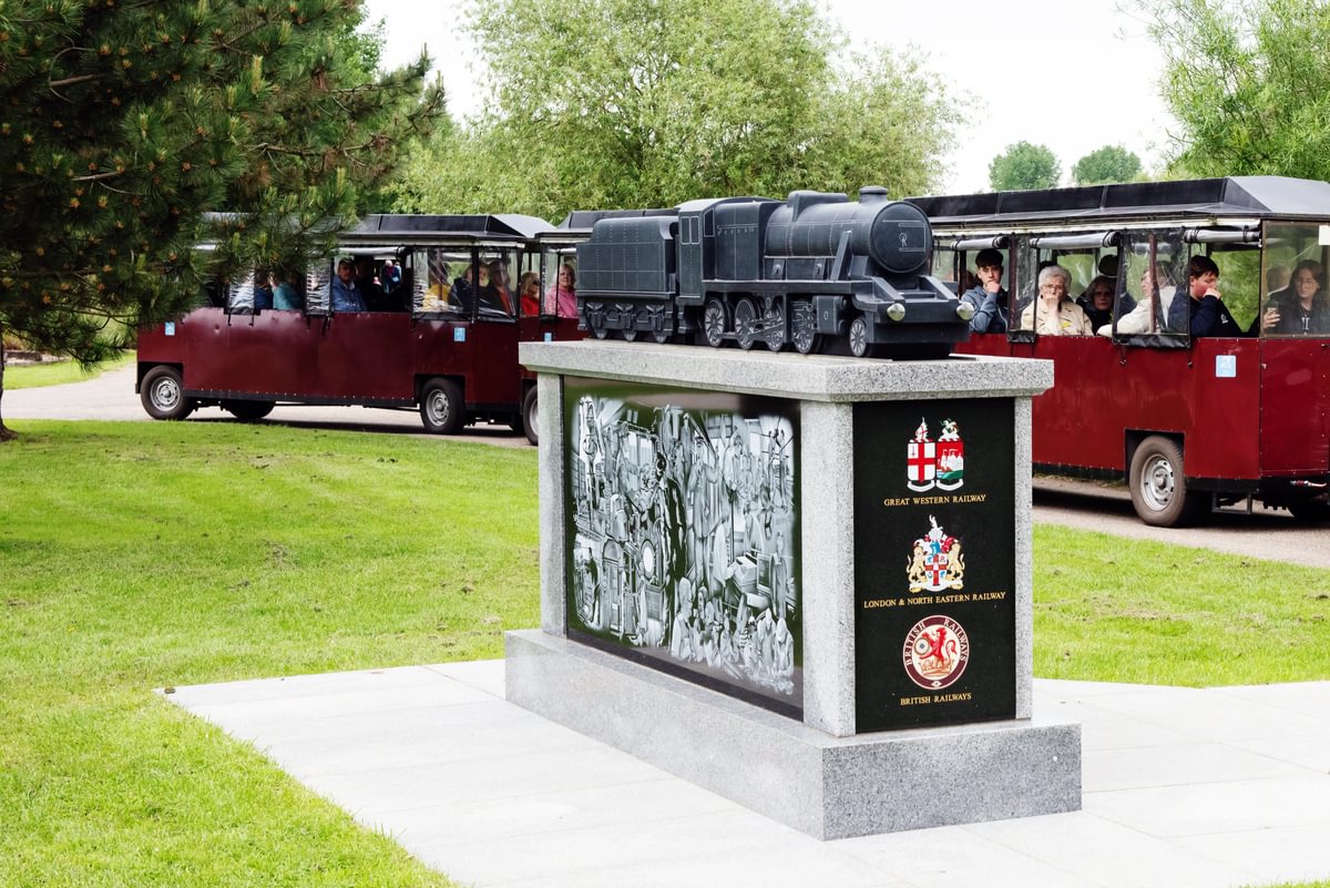 A red land train full of passengers travels past the Rail Industry Memorial, it is constructed from slate, and is distinctly shaped like a traditional locamotive.