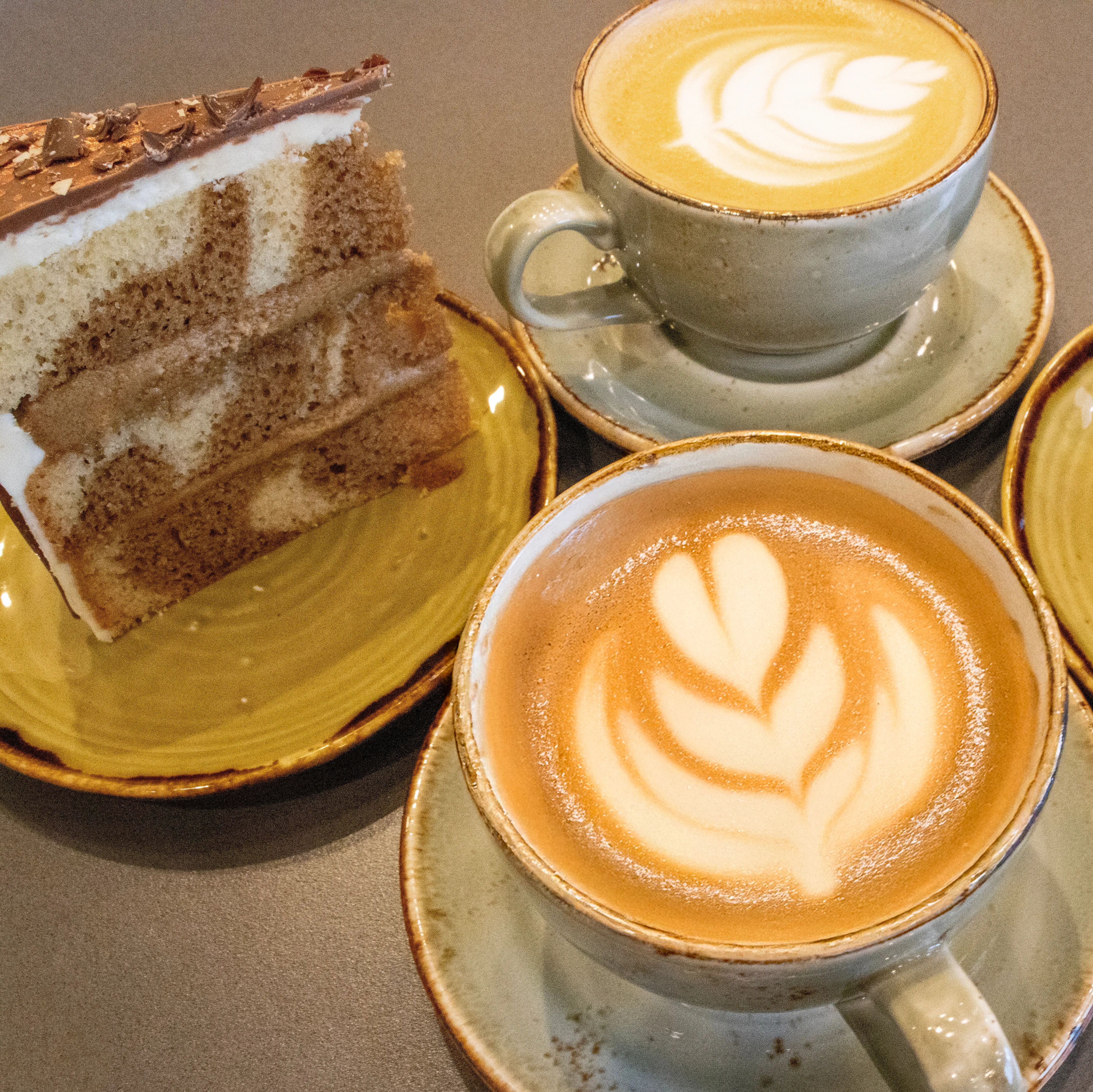 Barista fresh coffees in mugs at the Arboretum coffee shop, next to a slice of cake.