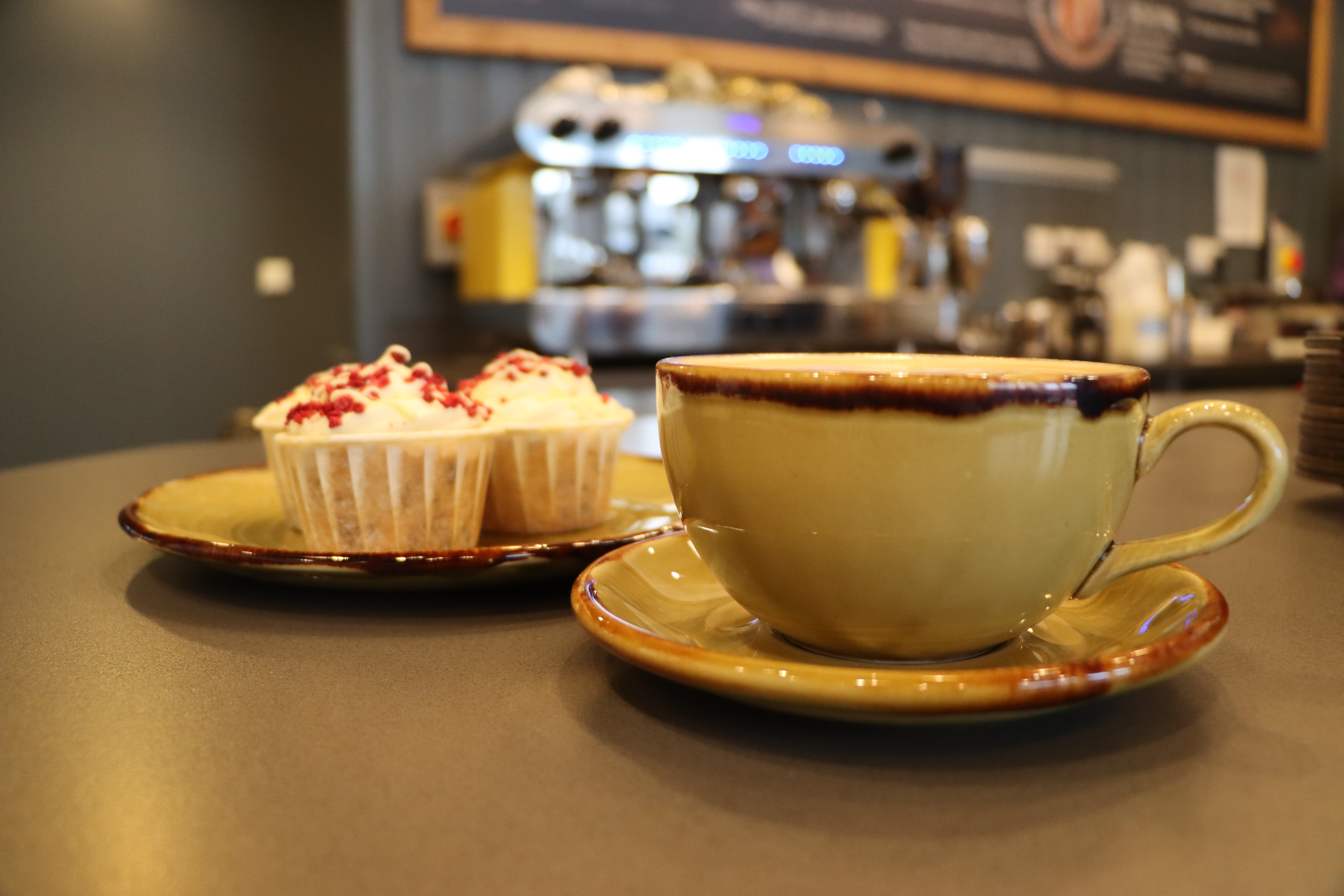 A cup of coffee in a large mug, sits in front of a plate of cupcakes within the arboretum coffee shop. The coffee machine is in the background.